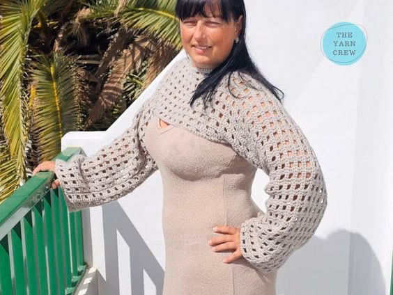 Three-quarter angled view of a woman standing by a balcony railing, modeling a beige crochet net mesh shrug with wide sleeves over a matching dress. Original design by Ariana of TheYarnCrew.