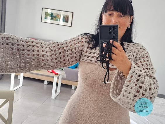 Mirror selfie of a woman holding her phone up, modeling a beige crochet net mesh shrug with wide, long sleeves over a matching dress. Original design by Ariana of TheYarnCrew.