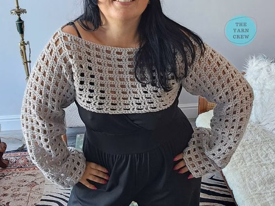 Front view of a woman standing with hands on her hips, modeling a beige crochet net mesh shrug with wide long sleeves over a black dress. Original design by Ariana of TheYarnCrew.