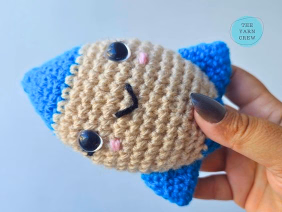 A close-up shot of a hand squeezing a small, soft crochet rocket toy to show how squishy it is. The rocket has a smiling face, a beige middle, and blue tips, set against a plain light background with a small logo in the corner. Original design by Ariana of TheYarnCrew.