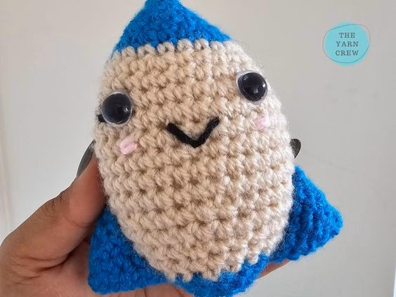 A close-up shot of a person holding a small, handmade crochet rocket toy. It has a beige middle, a bright blue top and bottom, and a friendly smiling face with shiny black eyes. The background is a plain wall, and there is a small logo in the corner. Original design by Ariana of TheYarnCrew.