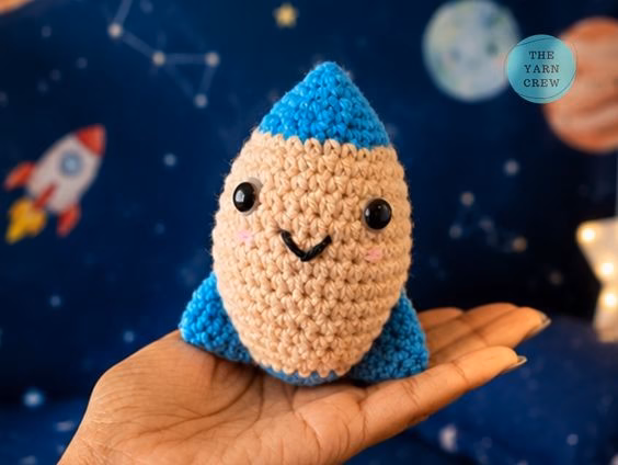 A person holding a small, handmade crochet rocket toy in their open hand. The rocket has a smiling face, a beige middle, and bright blue tips. The background is a blurry dark blue wall with a cartoon space theme, and there is a small logo in the top right corner. Original design by Ariana of TheYarnCrew.