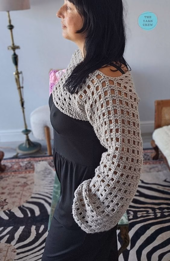 Side profile view of a woman modeling a beige crochet net mesh shrug with long, wide sleeves over a black dress. Original design by Ariana of TheYarnCrew.