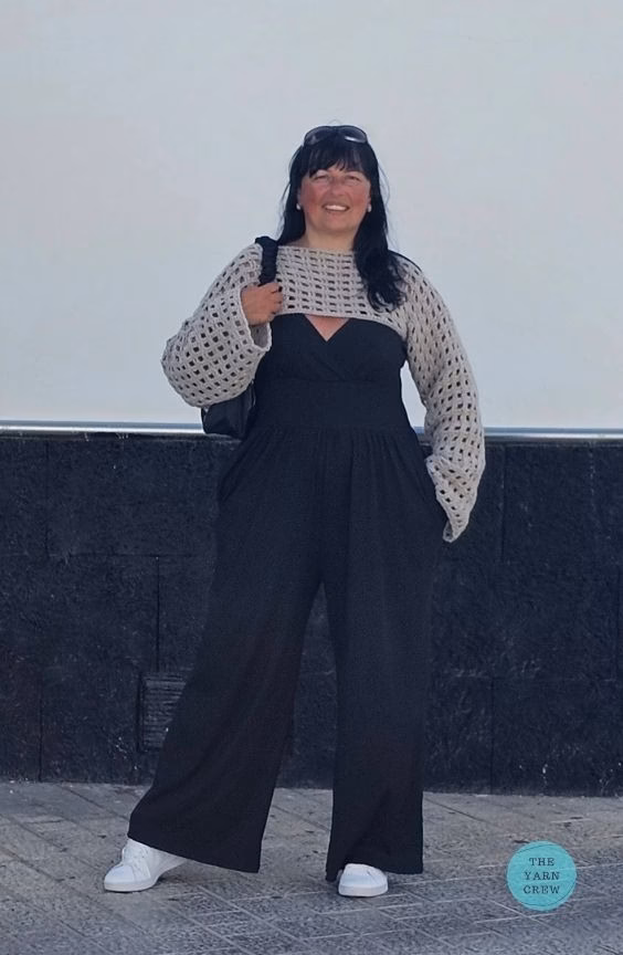 Full-length view of a woman standing outdoors, modeling a beige crochet net mesh shrug with wide sleeves over a black wide-leg jumpsuit and white sneakers. Original design by Ariana of TheYarnCrew.