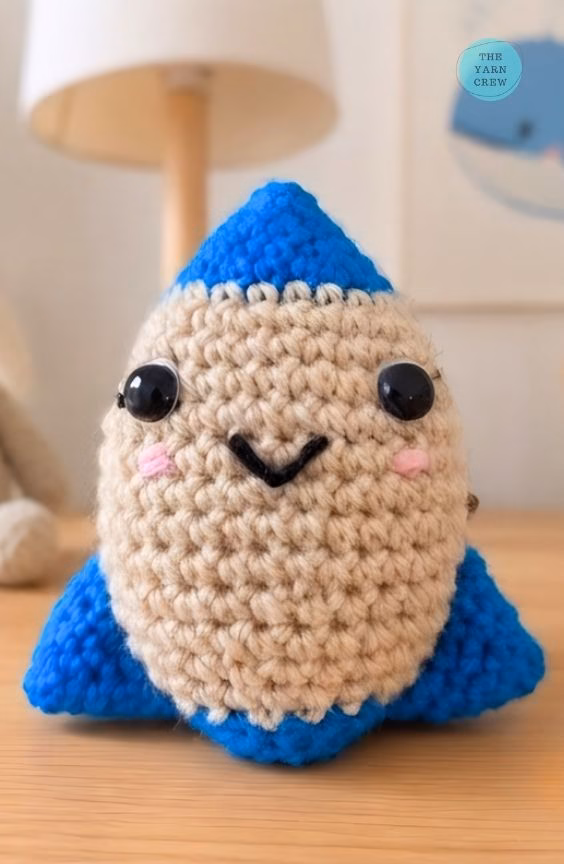 A close-up shot of a small, handmade crochet rocket toy sitting on a wooden table. It has a smiling face, a beige middle, and bright blue tips. The background is a blurry room with a lamp and a drawing on the wall, and there is a small logo in the top right corner. Original design by Ariana of TheYarnCrew.