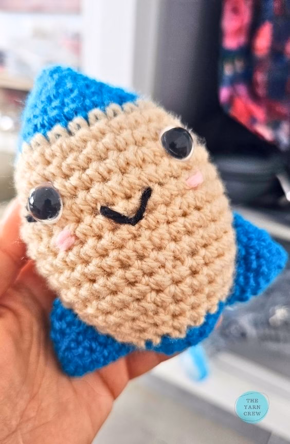 A close-up shot of a person holding the finished crochet rocket toy at a slight angle. It has a smiling face, a beige middle, and bright blue tips, with a blurry room in the background and a small logo in the corner. Original design by Ariana of TheYarnCrew.