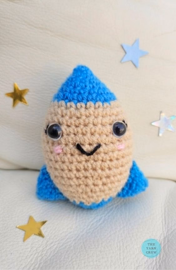 A small, handmade crochet rocket toy with a smiling face, beige middle, and blue tips. It sits on a white surface surrounded by a few little star stickers, with a small logo in the corner. Original design by Ariana of TheYarnCrew.