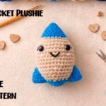 A cute, handmade crochet rocket toy with a friendly face, a beige middle, and a bright blue top and bottom. It sits on a table next to a wooden crochet hook, small golden scissors, and tiny wooden hearts. Text on the picture says "Rocket Plushie" and "Free Pattern." Original design by Ariana of TheYarnCrew.