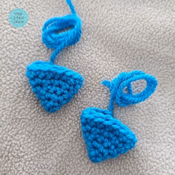 Two small, triangle-shaped pieces of bright blue crochet yarn lying on a soft grey surface. Each piece has a long loop of extra yarn attached to the top, with a small logo in the top left corner. Original design by Ariana of TheYarnCrew.