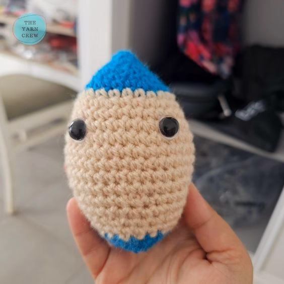 A person holding a small crochet rocket toy that is missing its smile and side fins. It has a beige body, black eyes, and blue tips, with a blurry room in the background and a small logo in the top left corner. Original design by Ariana of TheYarnCrew.