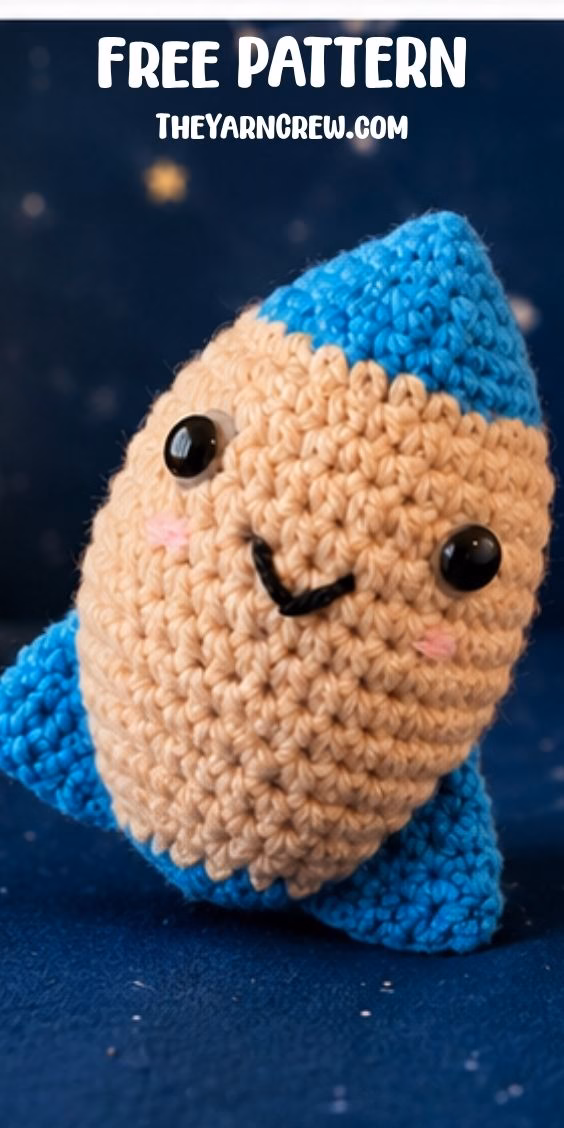 A close-up shot of a cute crochet rocket toy with a smiling face, beige middle, and blue tips. It stands on a dark blue surface, and text at the top reads "FREE PATTERN THEYARNCREW.com." Original design by Ariana of TheYarnCrew.