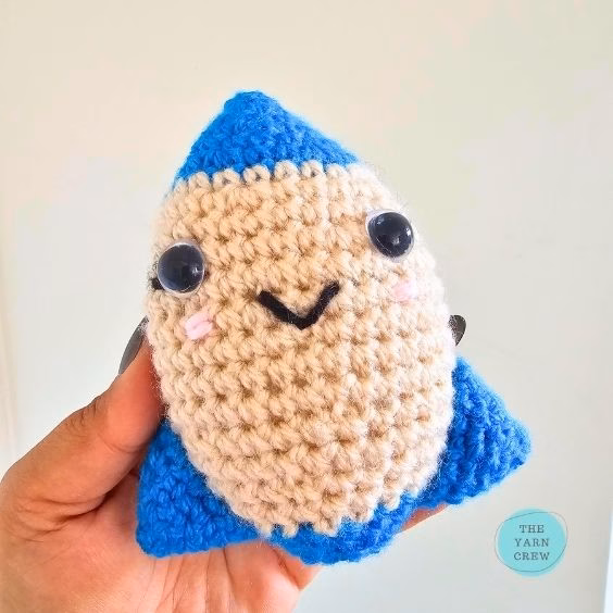 A person holding a small, handmade crochet rocket toy. It has a beige middle, a bright blue top and bottom, and a friendly smiling face with shiny black eyes. The background is a plain wall, and there is a small logo in the corner. Original design by Ariana of TheYarnCrew.