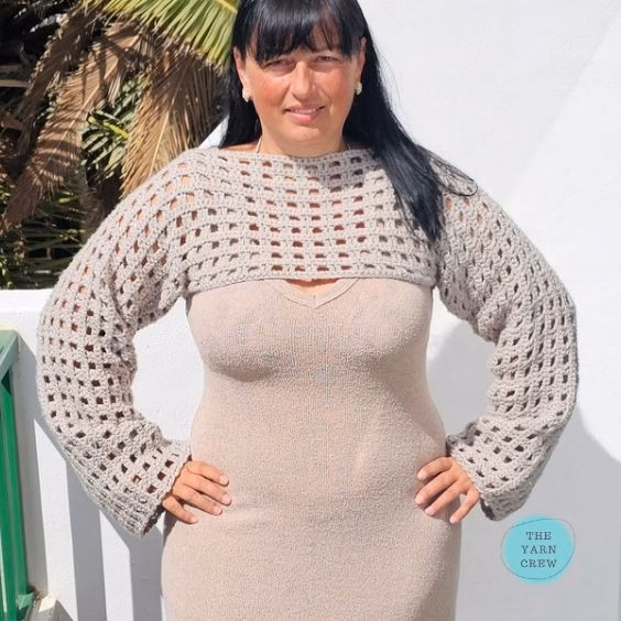 Woman standing with hands on her hips, modeling a beige crochet net mesh shrug with wide sleeves over a matching dress. Original design by Ariana of TheYarnCrew.