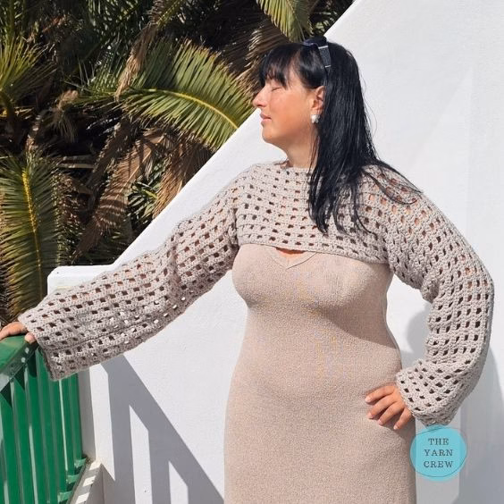 Side view profile of a woman looking away with her eyes closed, standing by a balcony railing and modeling a beige crochet net mesh shrug with wide, long sleeves over a matching dress. Original design by Ariana of TheYarnCrew.