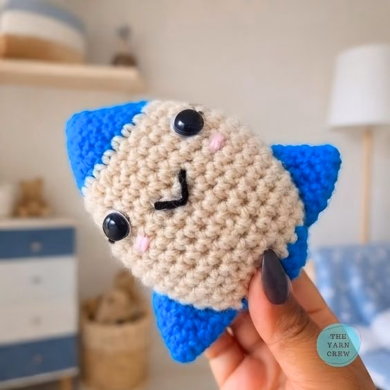 A close-up shot of a person holding the finished crochet rocket toy tilted sideways. It has a smiling face, a beige middle, and bright blue tips, with a blurry bedroom in the background and a small logo in the corner. Original design by Ariana of TheYarnCrew.
