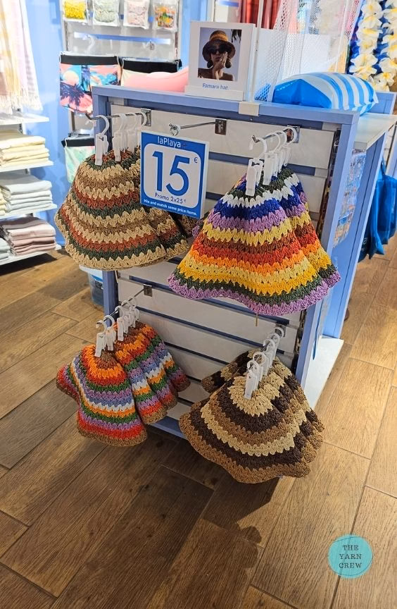 A display stand of crocheted bucket hats in various striped color combinations hanging in a retail store. Original design created by Ariana Wimsett at TheYarnCrew.com.