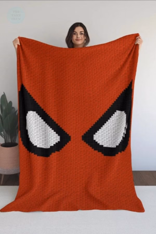 A woman holding up a large, solid red c2c crochet blanket featuring giant Spider-Man eyes made of white blocks with thick black borders. This is a crochet roundup from https://theyarncrew.com/.