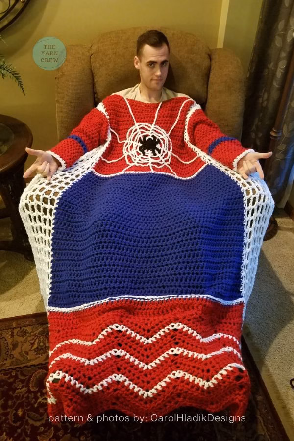 Image 1: A man sitting in an armchair wearing a wearable Spider-Man themed crochet blanket with sleeves. The top section is red with a white crochet spiderweb and black spider applique, the middle is solid blue, and the bottom features red and white chevron stripes. This is a crochet roundup from https://theyarncrew.com/.